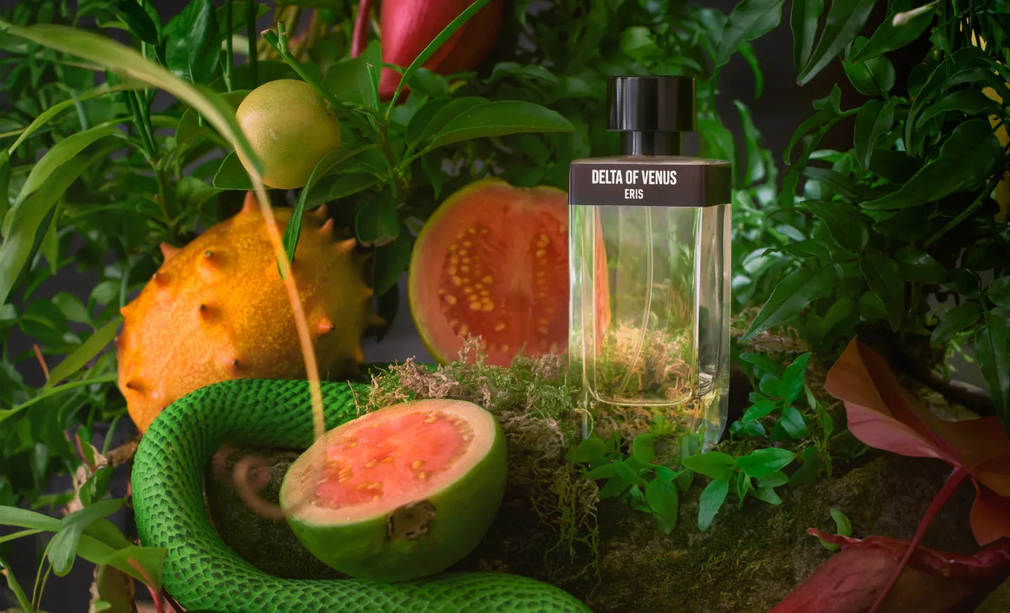 A perfume bottle labeled Delta of Venus by Eris is placed among lush greenery and halved exotic fruits. The scene is bright and natural, featuring various shades of green, pink, and orange, conveying an earthy, fresh vibe.