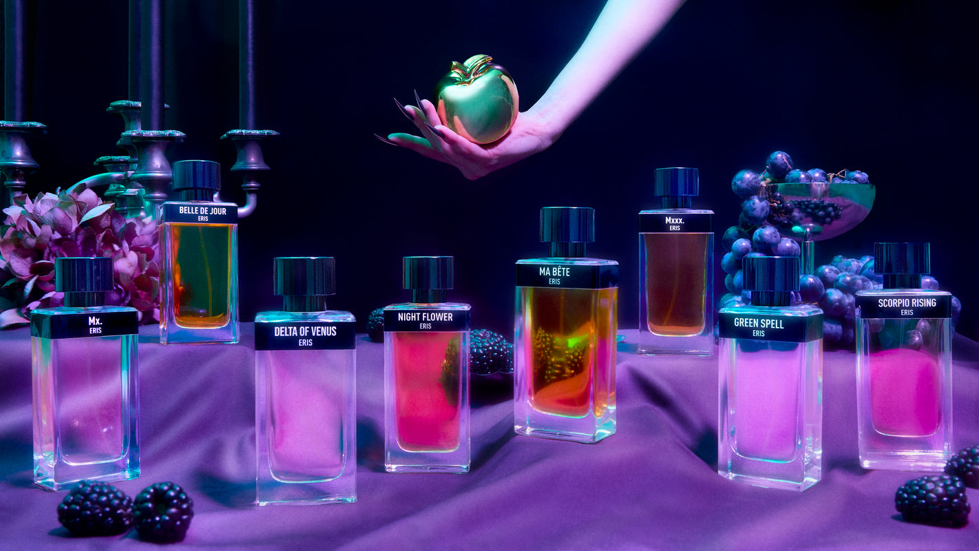 Perfume bottles from Eris Parfums sit on a purple tablecloth, filled with colorful liquids and diverse labels. A hand holds a golden apple above them amidst candles and fruit, creating an enchanting backdrop under dramatic lighting.